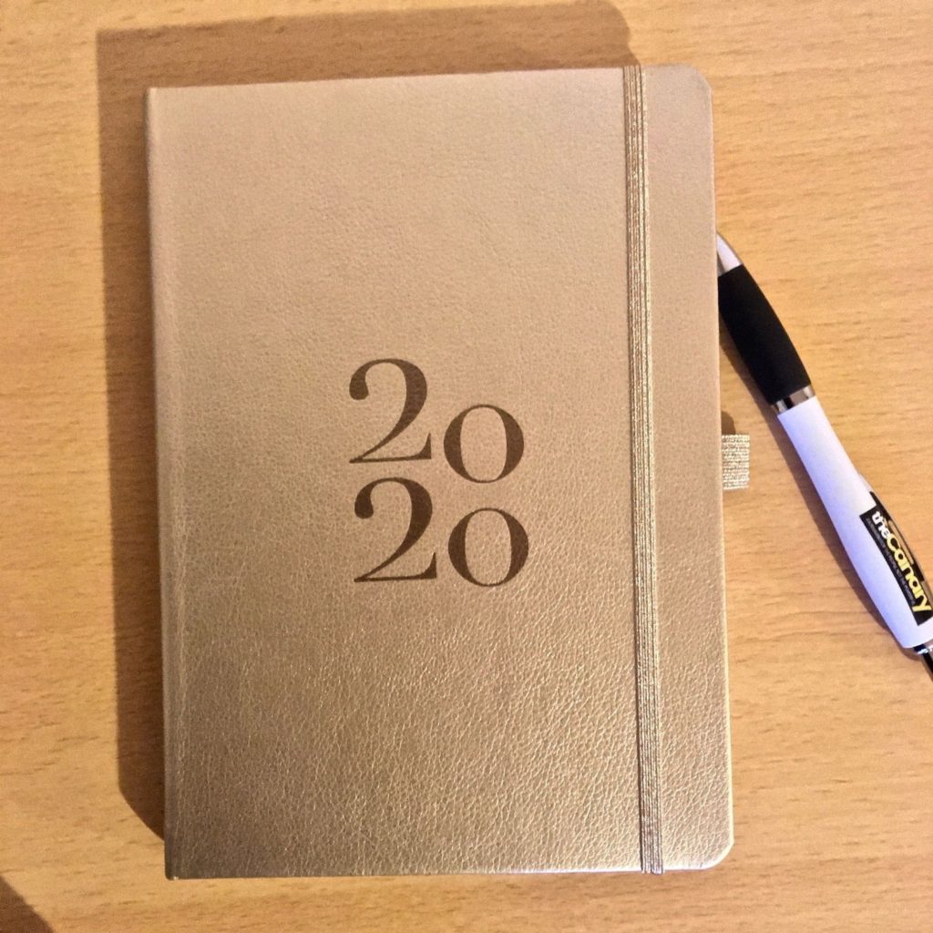 Photo of A5 size planner with gold-colour binding, and ‘2020’ embossed on front cover. Next to it is branded pen from The Canary website.
